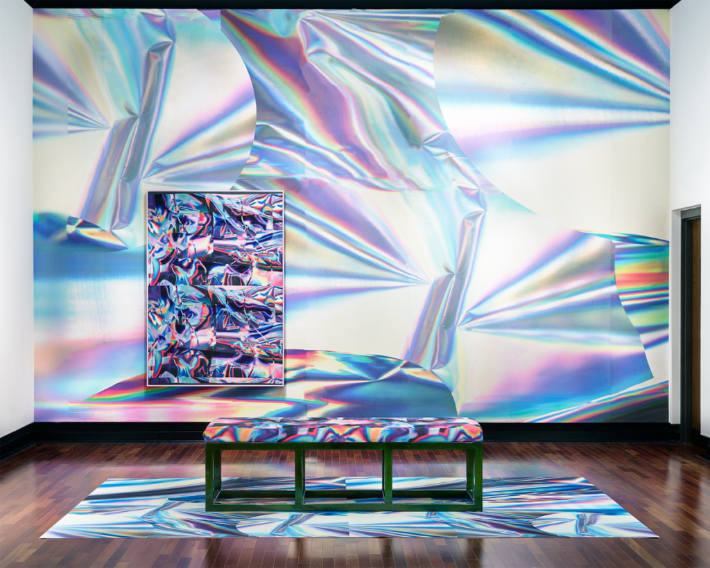 Installation view of rainbow and chrome abstract designs projected onto a wall in front of a bench. On the wall hangs an artwork with a similar design.