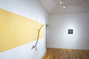Art exhibition featuring a bare tree branch attached to a white wall using thin horizontal yellow strips; the top is fully wrapped, bottom exposed.