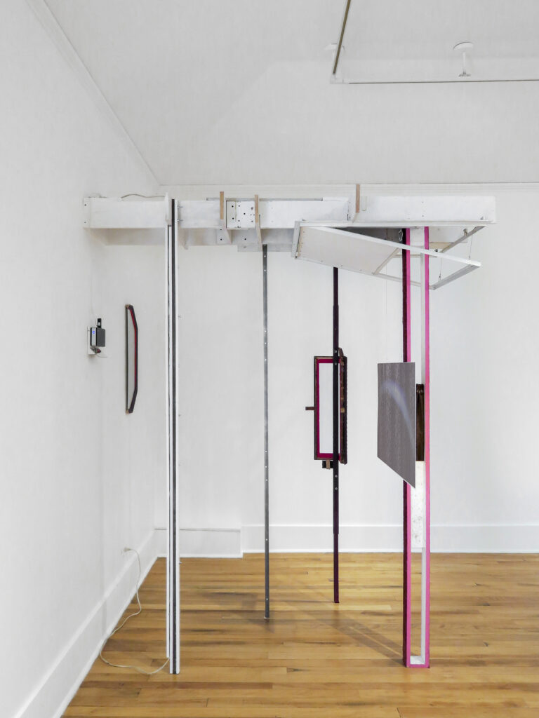 A gallery space with poles reaching from the floor to the ceiling, each one placed in front of the other, with a vent-like structure on the ceiling.