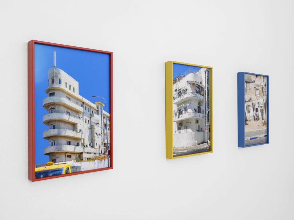 Three framed photographs line a white gallery wall, the first in a red frame, the next two in a yellow and blue frame, each holding an image of multi-tiered buildings and city life.