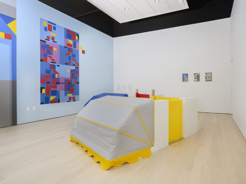 Installation view of a car-like sculpture with the front covered in grey material, and the back made of yellow and white rectangles. The wall shows a brightly colored geometric artwork.