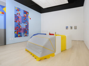 Installation view of a car-like sculpture with the front covered in grey material, and the back made of yellow and white rectangles. The wall shows a brightly colored geometric artwork.