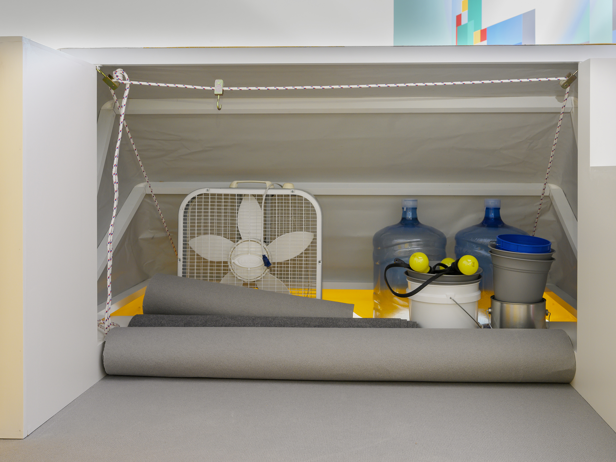 A gray makeshift tent lies on the exhibition floor holding a white fan, two empty jugs of water, a collection of plastic buckets, a string of yellow lightbulbs, and three rolls of gray paper. A white rope holds the structure together.