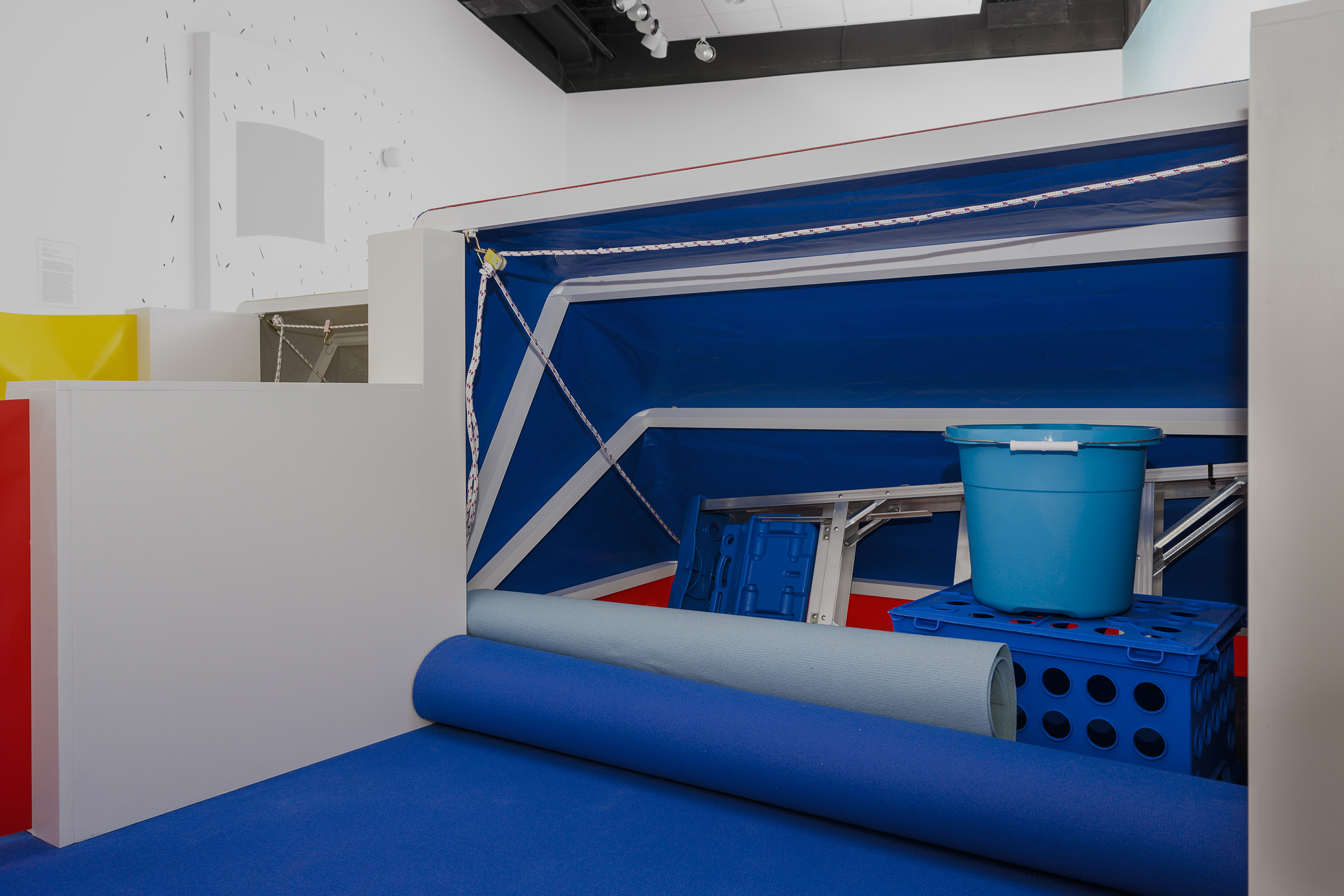 A blue makeshift tent lies on the exhibition floor holding a ladder, blue bucket, a crate, and two rolls of blue paper inside of it. A white rope holds the structure together.