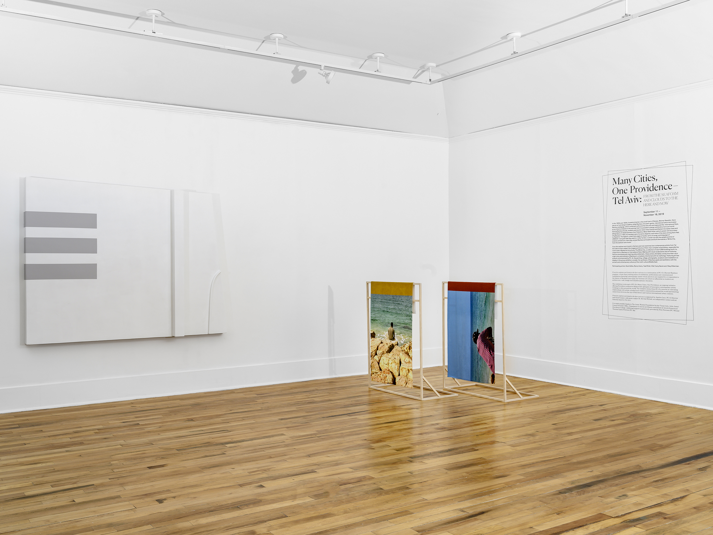 Installation view of a large white rectangular artwork with three grey horizontal stripes. To the right, two large photographs are displayed on wooden stands. Behind, the wall reads: Many Cities, One Providence - Tel Aviv.