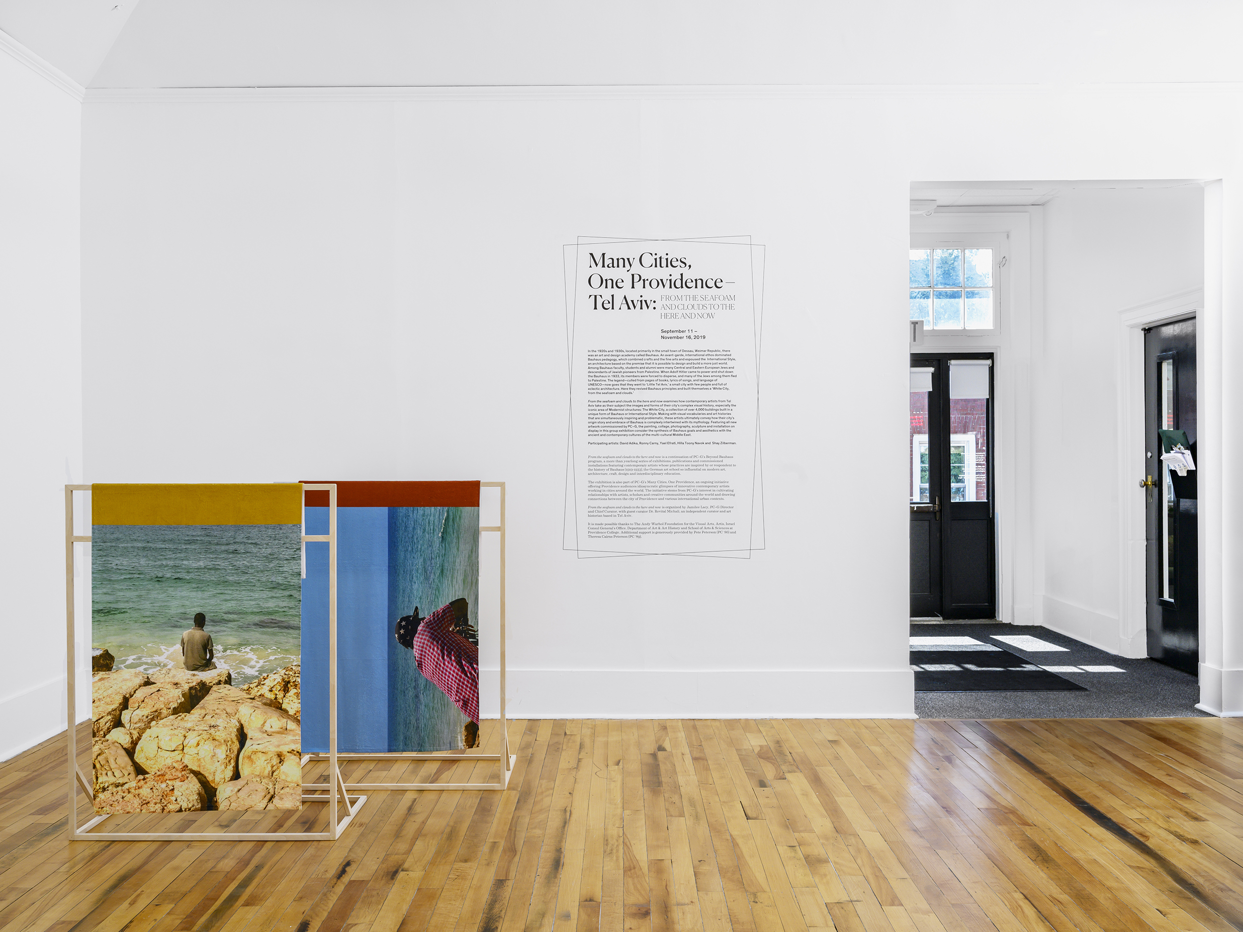 Installation view of two large photographs positioned vertically on wooden stands on the floor. Both include a figure sitting by the ocean. Behind, the wall reads: Many Cities, One Providence - Tel Aviv.