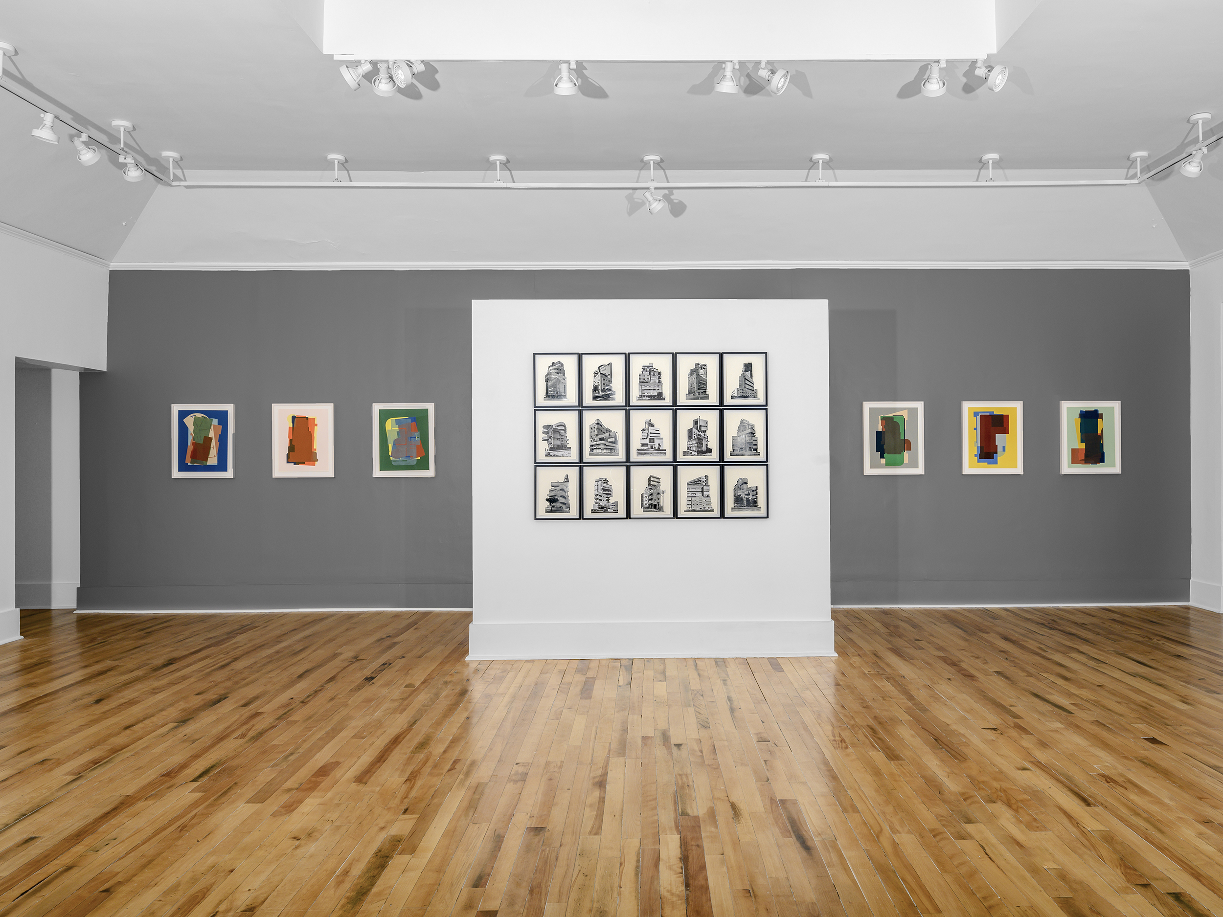 Installation view of a square white wall with a 3x5 array of black and white architectural photographs. Behind, a dark grey wall with three abstract artworks on either side.
