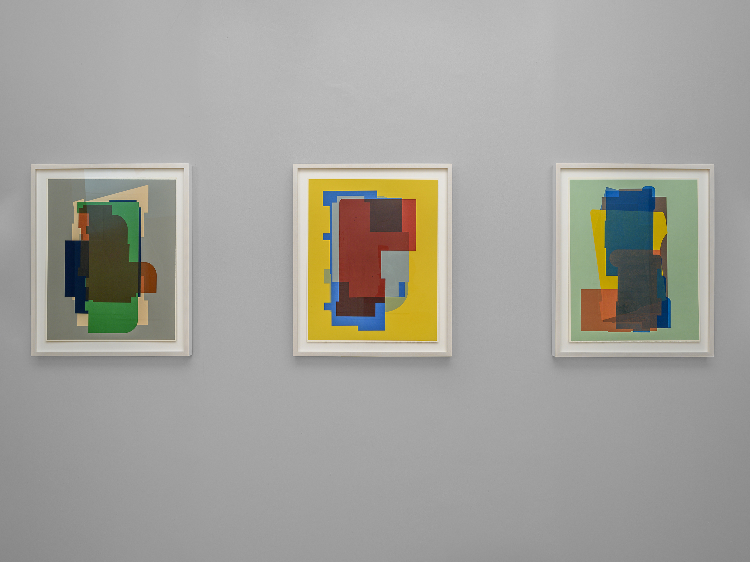 Three framed artworks on a dark grey wall. Each depicts overlapping geometric shapes in the center of a solid colored background, from left to right, grey, yellow, then light green.