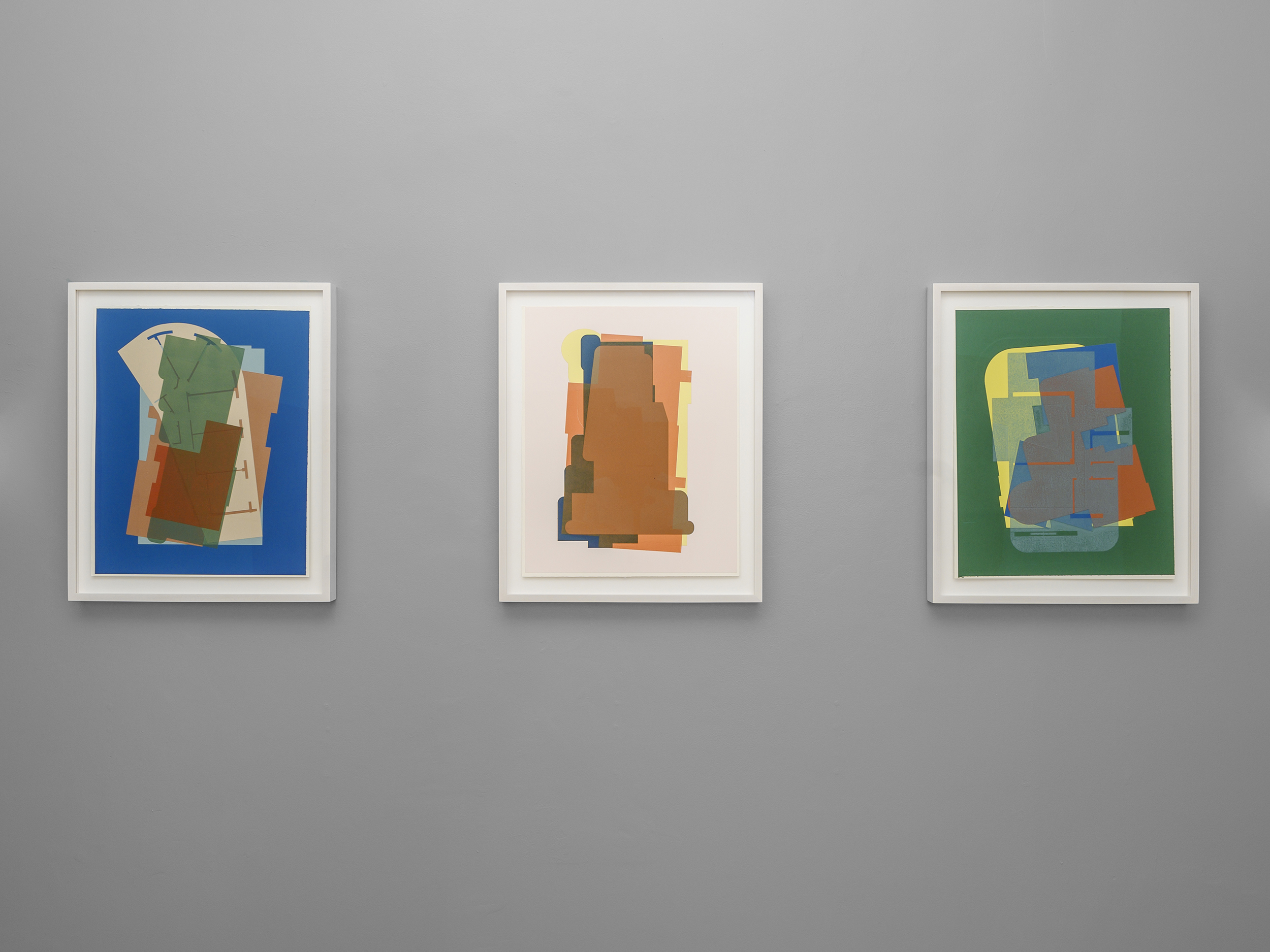 Three framed artworks on a dark grey wall. Each depicts overlapping geometric shapes in the center of a solid colored background, from left to right, blue, pink, then green.