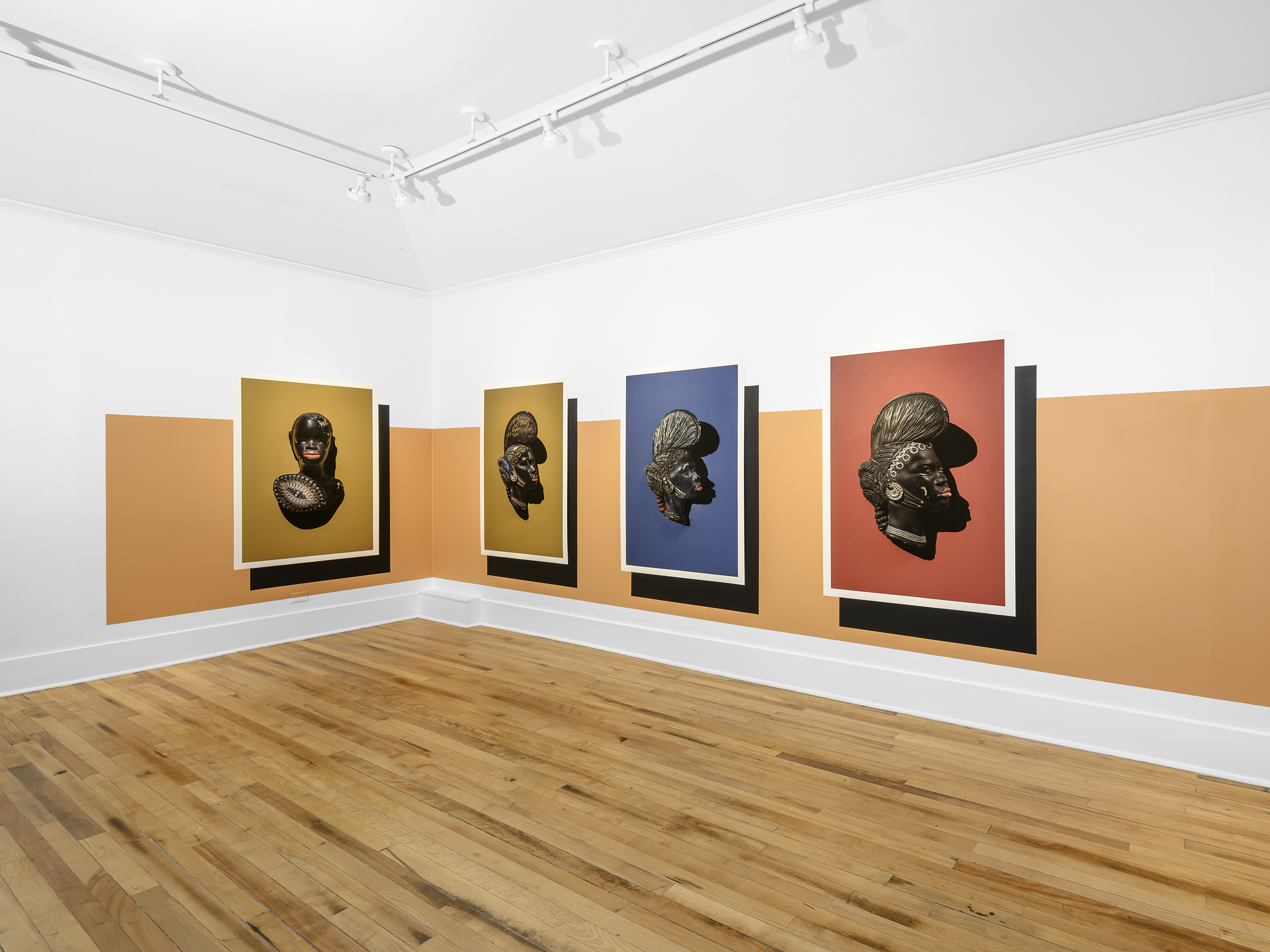 Installation view of a horizontal orange stripe spanning across two walls behind four artworks. Each artwork shows a different angle of a dark-skinned figure's head with a colorful solid background.