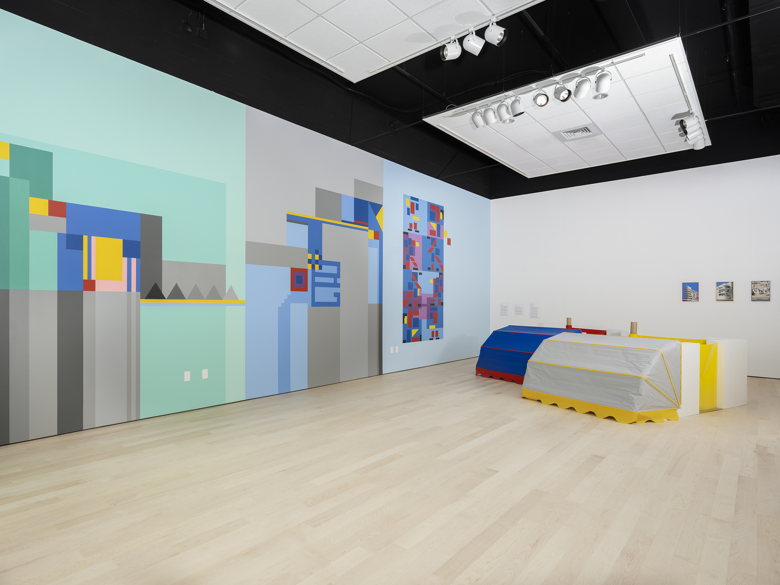 Installation view of a wall divided into three different rectangles of color, each further decorated with colorful geometric shapes. Two large sculptures similar to cars/carriages sit on the floor.