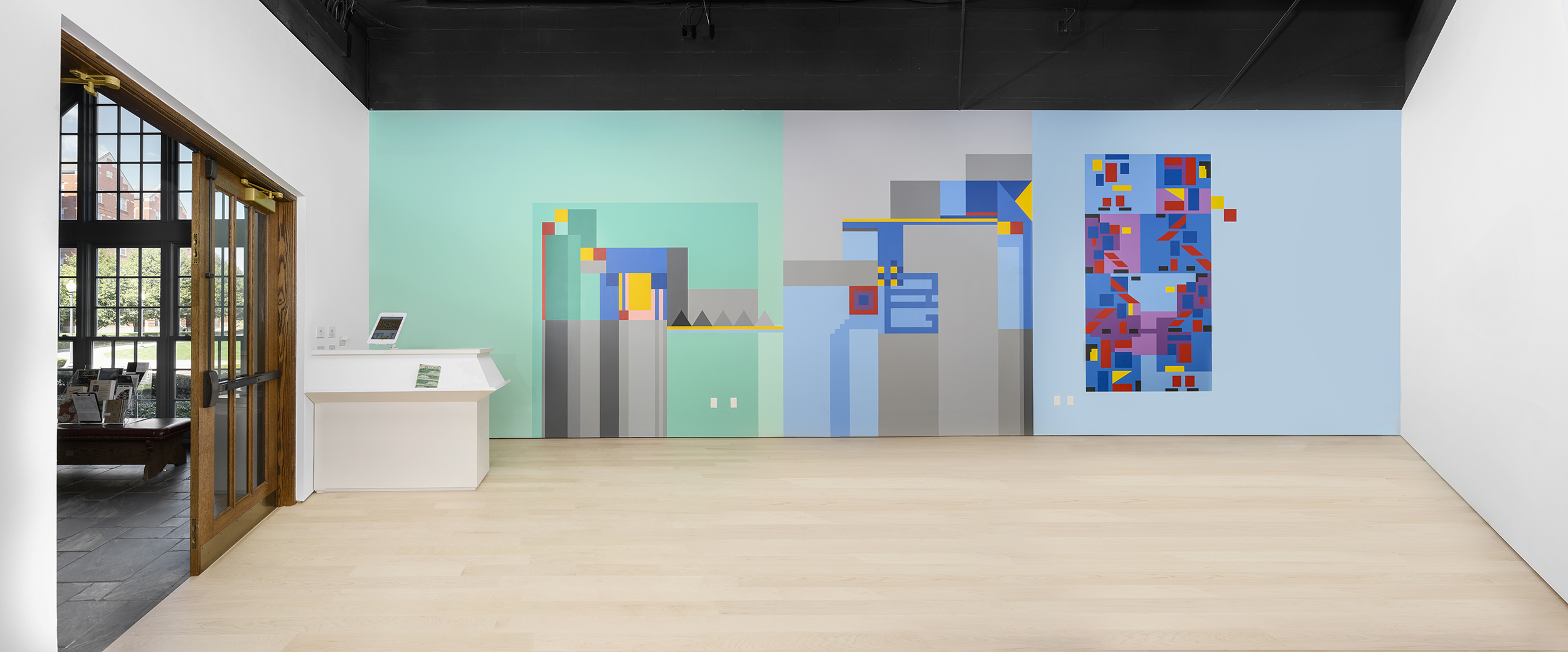 Installation view facing a large wall in an empty gallery. The wall is separated into three rectangles - green, grey, and blue. Each is further decorated by colorful abstract geometric shapes.