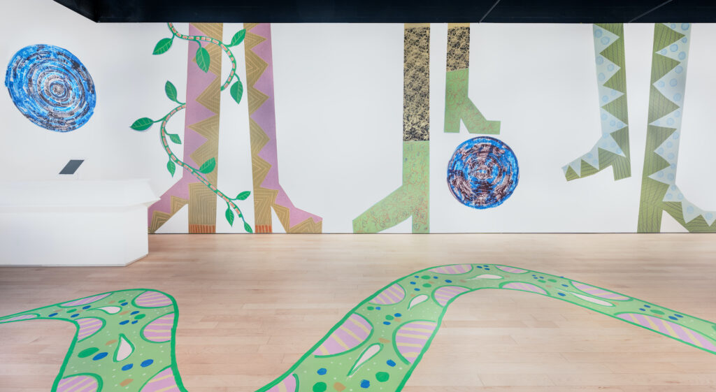 A gallery space with large cartoonish legs and feet painted on the wall in green, purple, and blue. A green, snake-like figure is painted on the gallery floor, winding up and down.