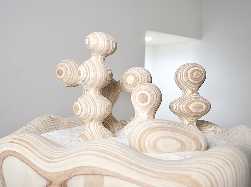 A wooden and sand sculpture in blob-like formations with different patterned bands sits in a white-wall gallery space.