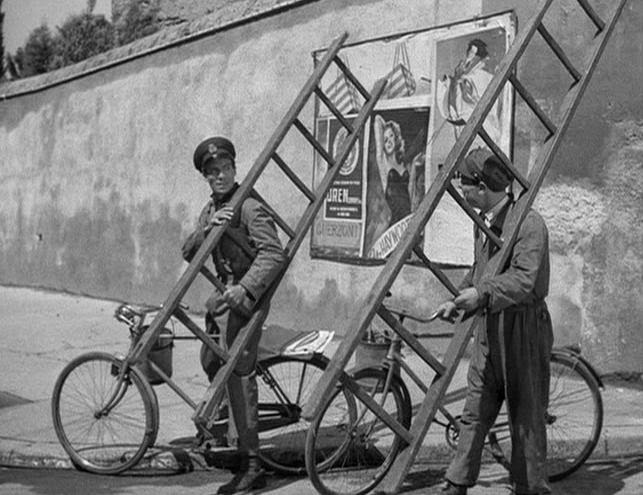 A black and white photograph of two men in military gear riding bicycles. They are both carrying tall wooden ladders on their shoulders and rest in front of a large concrete wall.