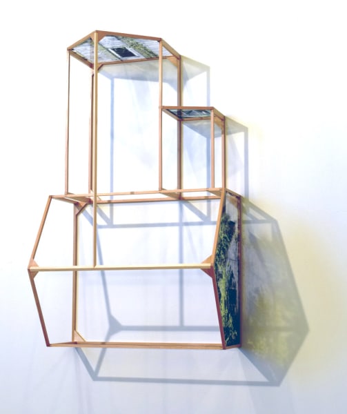 Slightly angled view of a wooden house frame mounted to a white wall. Three walls of the unusual structure are indicated by transparent photographs which cast dramatic shadows on the wall.