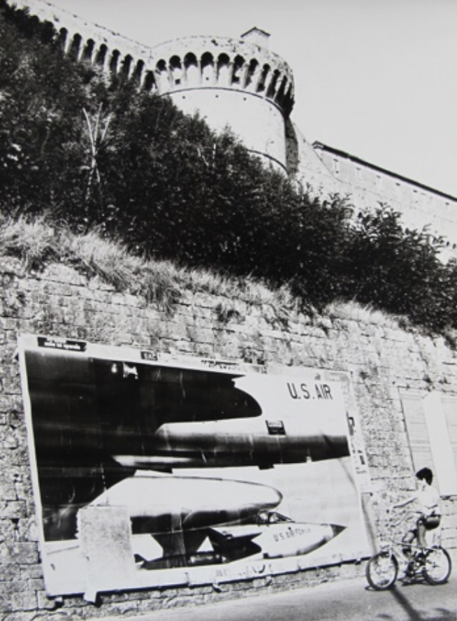 A brick wall holding a large poster depicting multiple planes with U.S. Air written on them. A young boy rides his bike in front of the poster, studying its content.