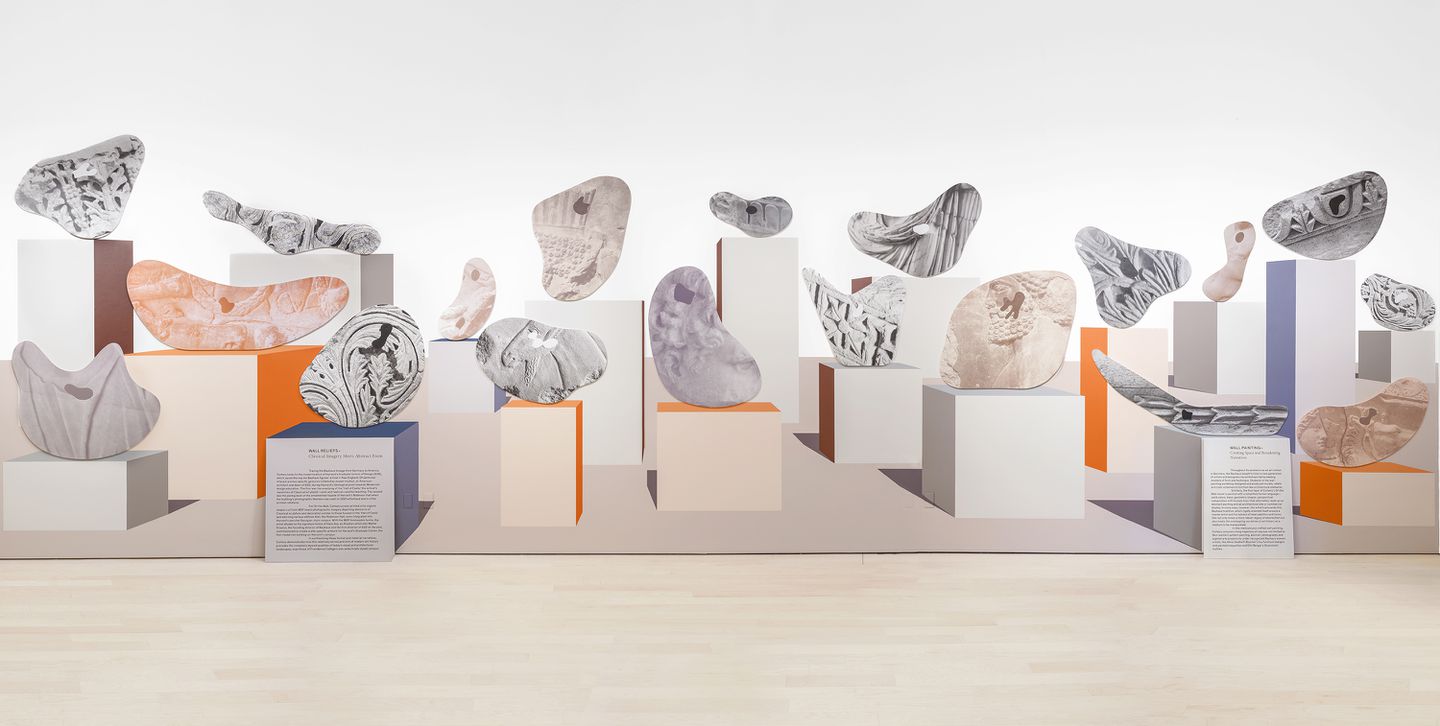 View of a white gallery wall with a panorama print on it depicting 19 cellular-shaped, multipatterned objects on different sized pedestals.
