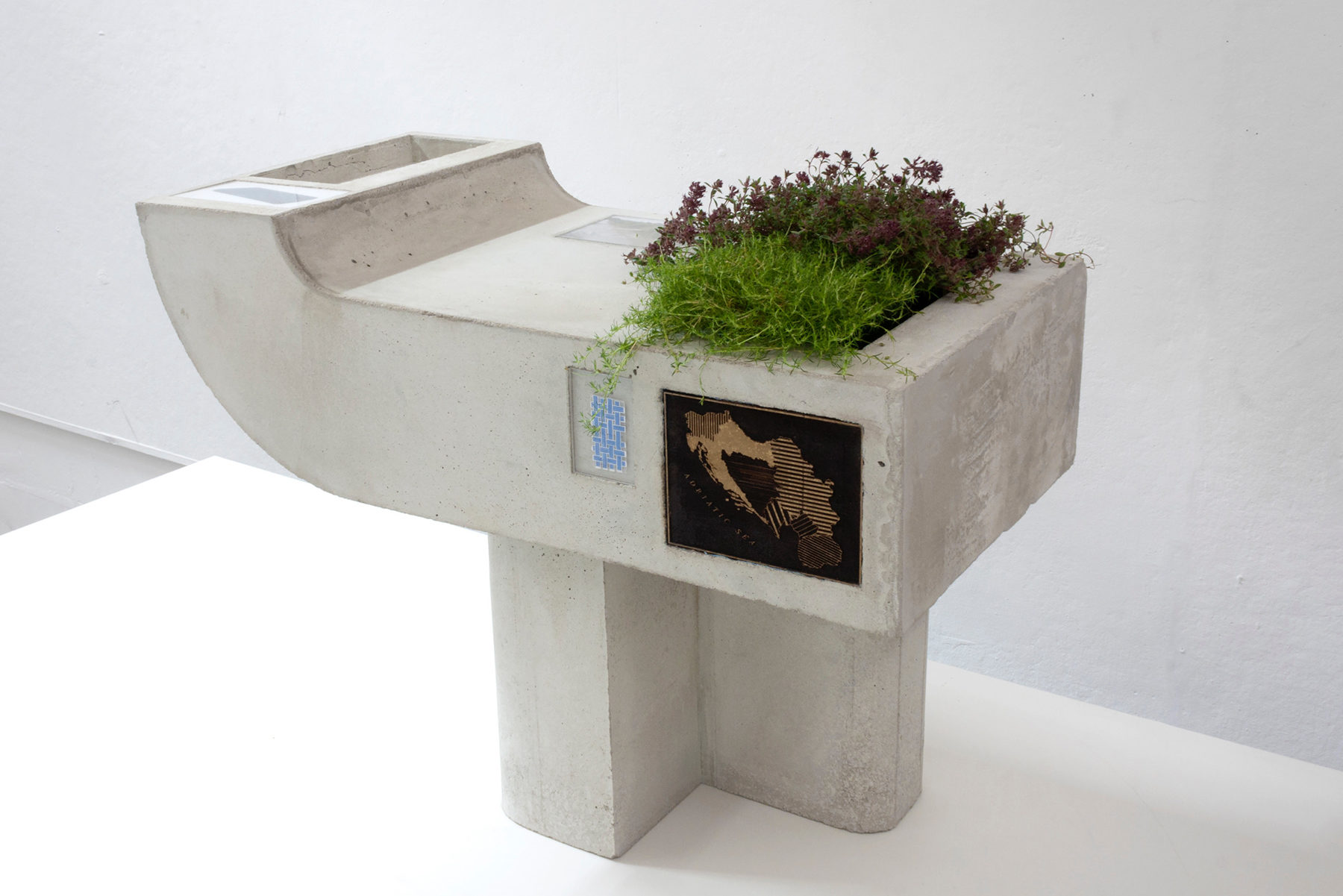A rectangle cement sculpture holding a thyme plant on the right side of it, with an L-shaped pedestal as a support.
