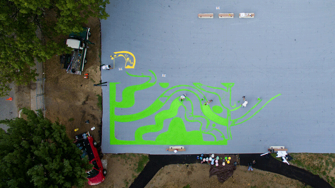 Bird's-eye view of a basketball court being painted with several green patterns and lines in the bottom right hand corner. The rest of the court remains a gray-blue color.