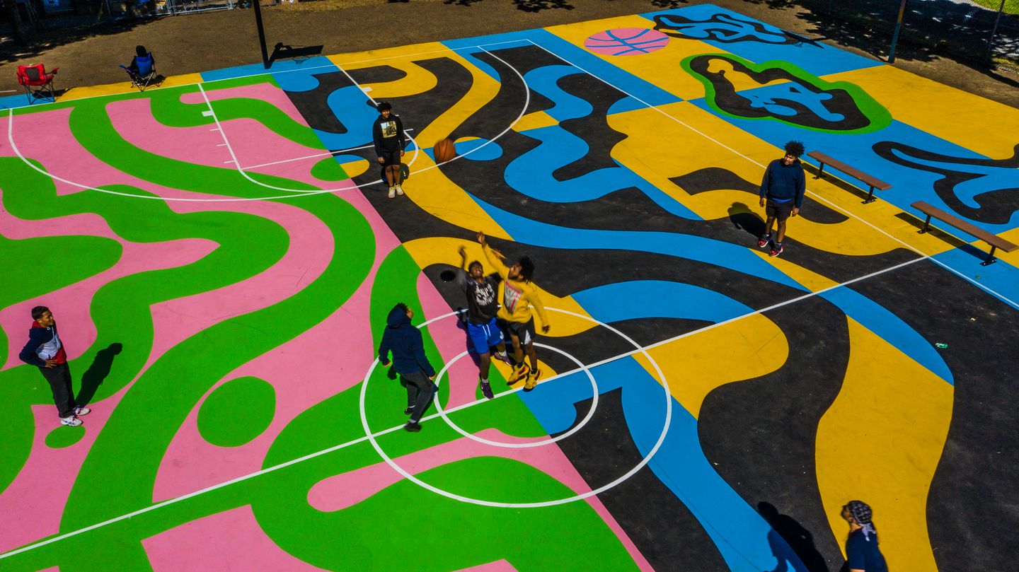 Overhead view of a basketball court with multicolor waves and lines decorating the surface. Seven people are standing on the court playing with a basketball being tossed in the air.