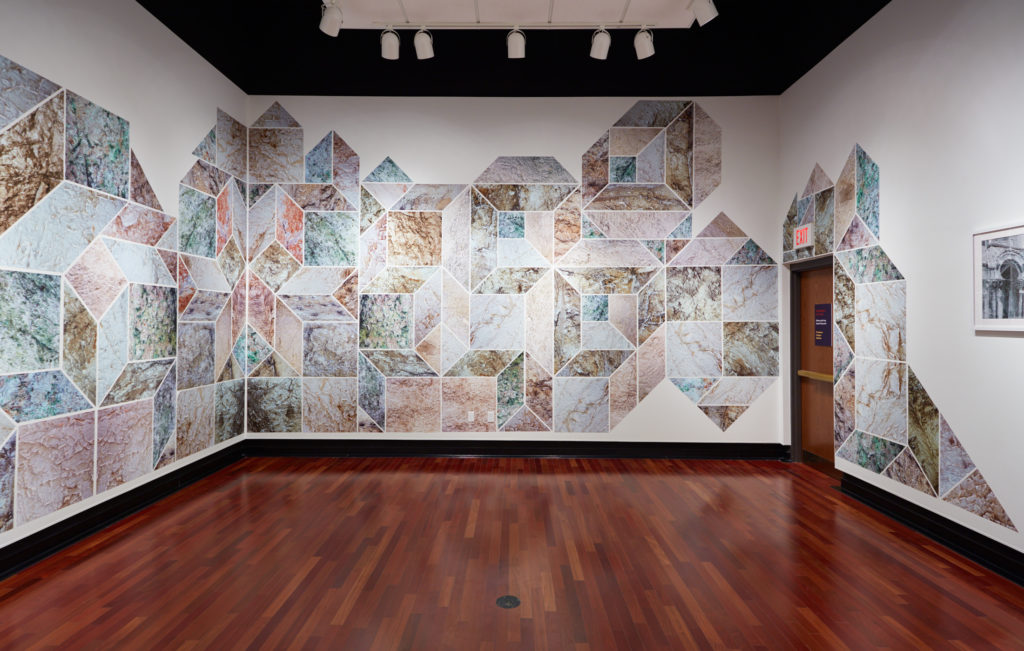 Installation view featuring two adjacent white walls connected through a huge design made up of interlocking green, orange, and beige geometric shapes, including squares, parallelograms, and diamonds.