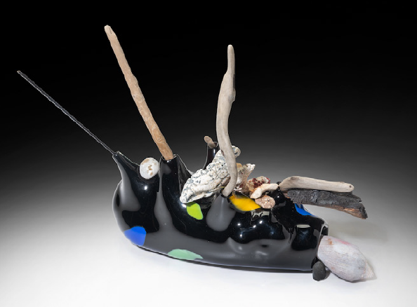 A black abstract sculpture with wooden protrusions on a black and white ombré background. The sculpture has colorful dots and organic matter placed on top.