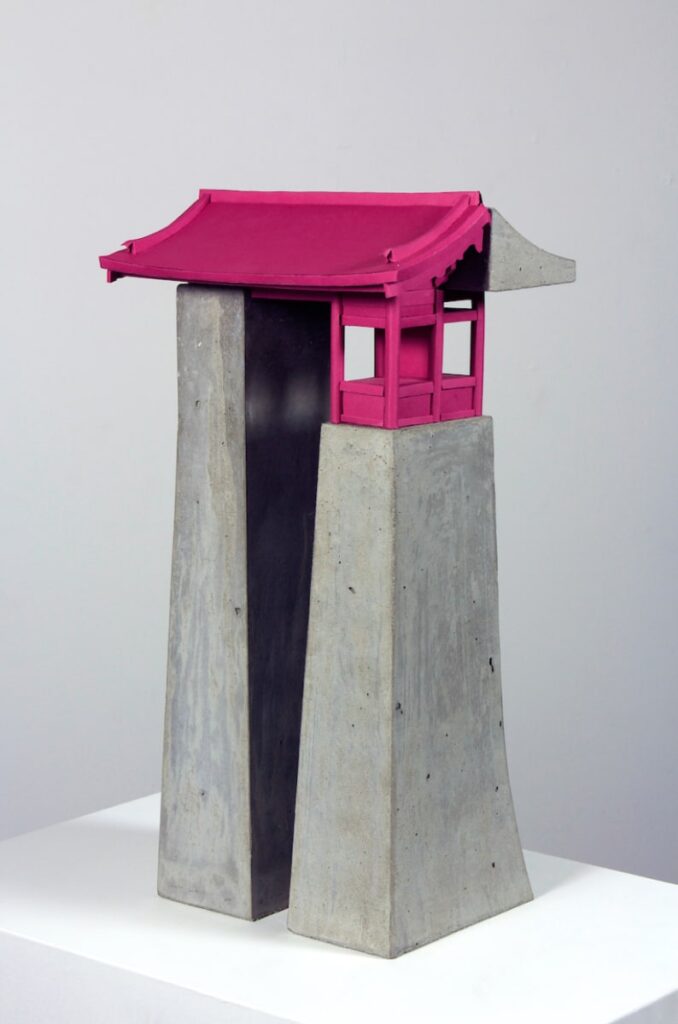 A sculpture of a vertical architectural form mostly made of grey concrete. Half of the roof and the front facade are hot pink.
