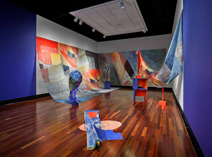 Installation view of large expanses of fabric filled with brightly colored shapes covering two walls. To the right, orange and blue structures uphold more fabric with similar colors.