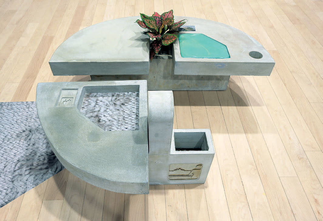 A grey sculpture with three pieces creating three-quarters of a circle. A plant sits between the top two pieces. To the left on the floor is an abstract grey pattern.