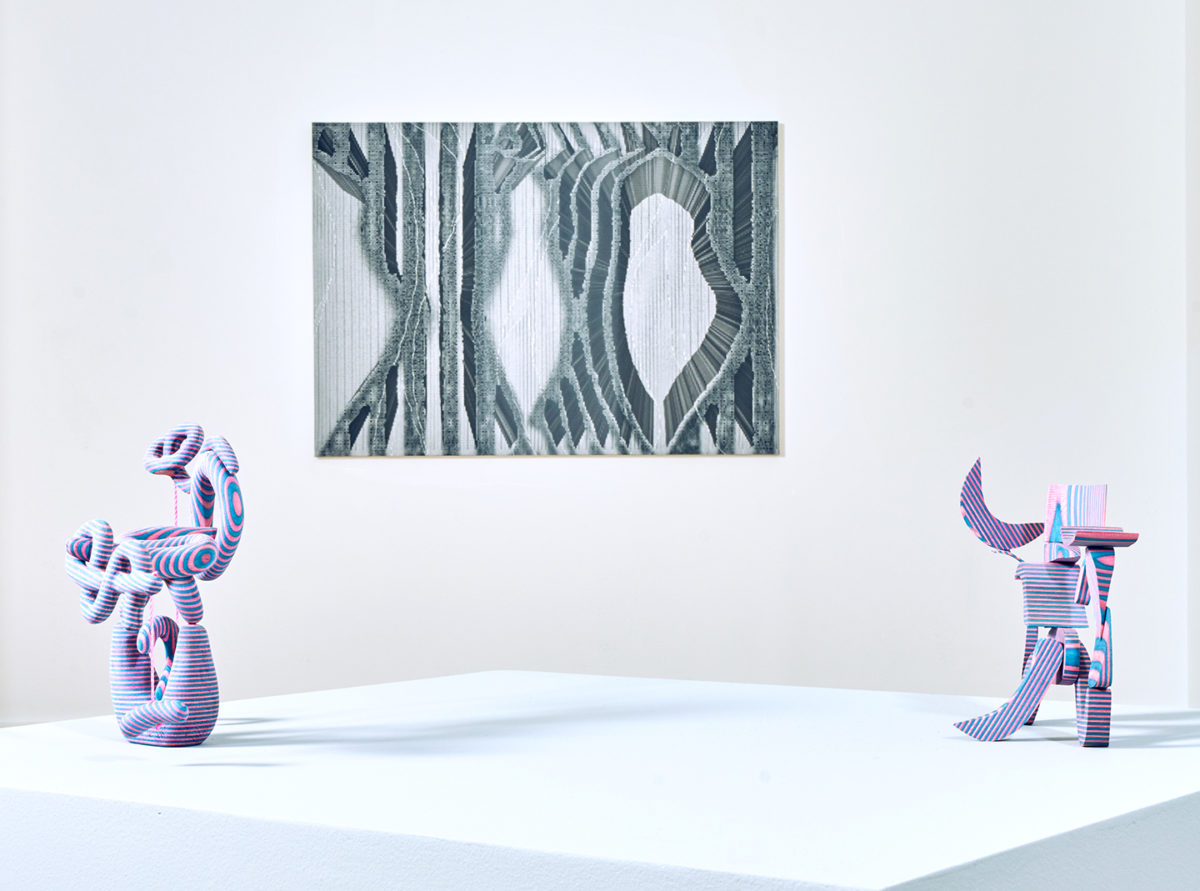 Two pink and blue striped abstract sculptures sit on a white box. On the wall behind them is an abstract black and white artwork of curving lines.