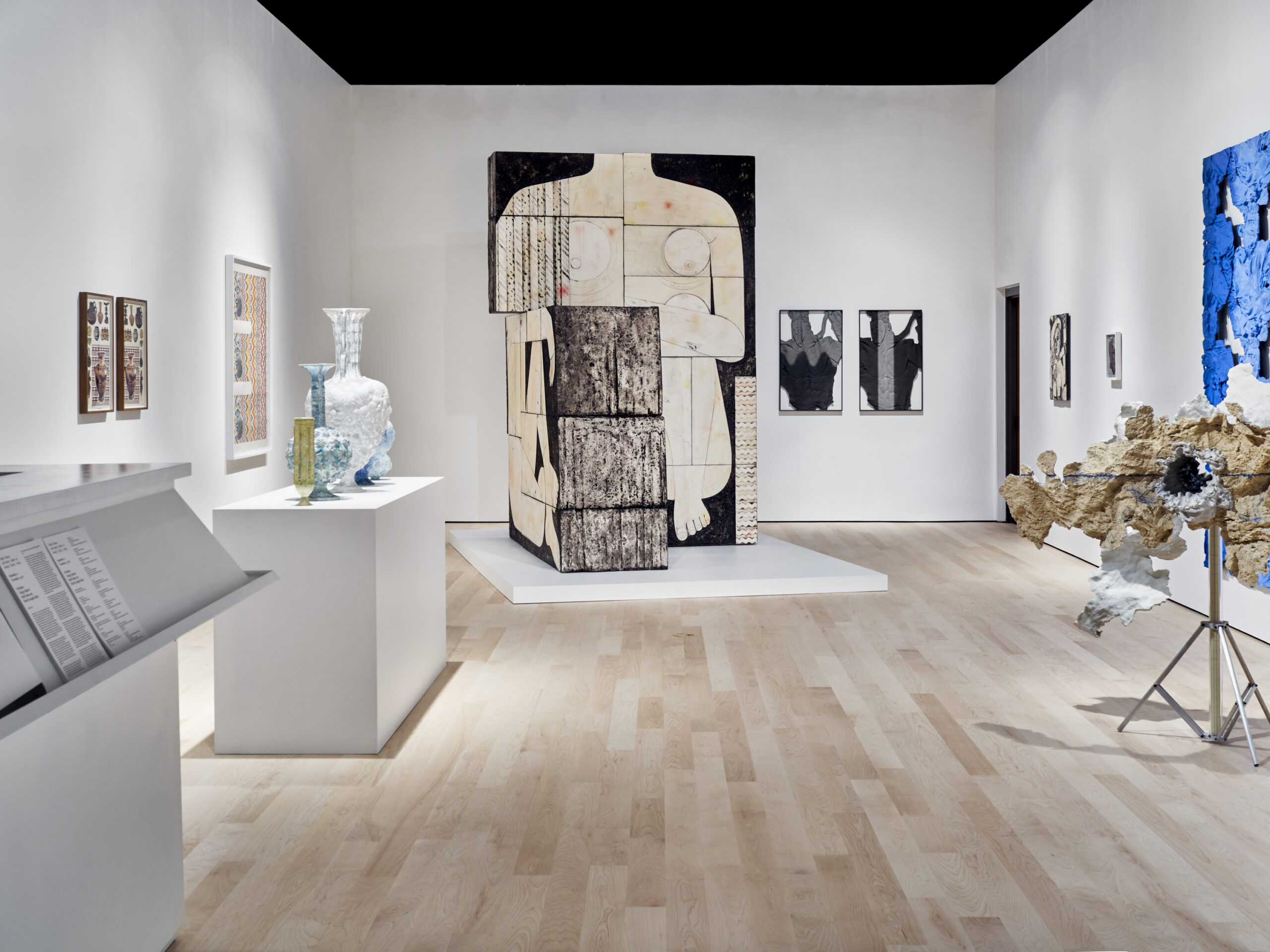 Installation view of an Art exhibition featuring ceramic vessels on a white pedestal, a headless, white-skinned, naked female figure drawn on large boxes, framed prints on walls, and organic textured sculptures.