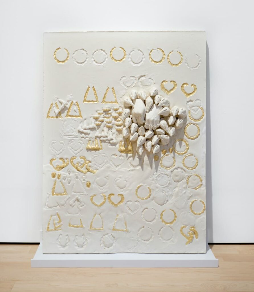 Textured white panel featuring gold-outlined imprints of hearts, circles, and triangles. At its center, a sculpture of white fingers, fists, and beaks protrudes outward in dense formation.
