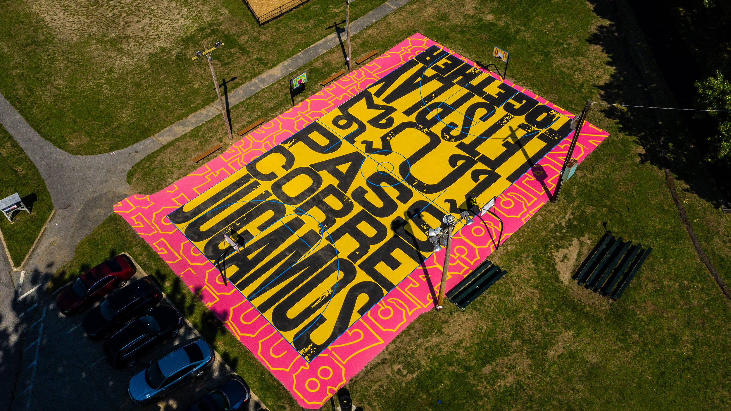 A bird's eye view of a yellow basketball court with a pink border around it. On the court written in black text are the words PASS CORRE JUGAMOS and LET'S PLAY TOGETHER.