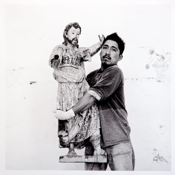 A man stands holding an old, damaged statue. He looks directly at the camera with a serious expression. The statue is missing parts and shows signs of age and wear.
