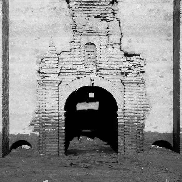 A crumbling brick arch stands tall with light peeking through the tunnel behind it, in black and white.