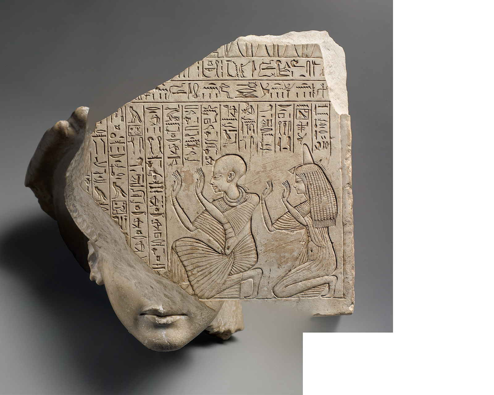 Beige stone tablet with ancient Egyptian carvings of people and symbols, merged with a realistic human face at the viewer's bottom left, set against a gray background.