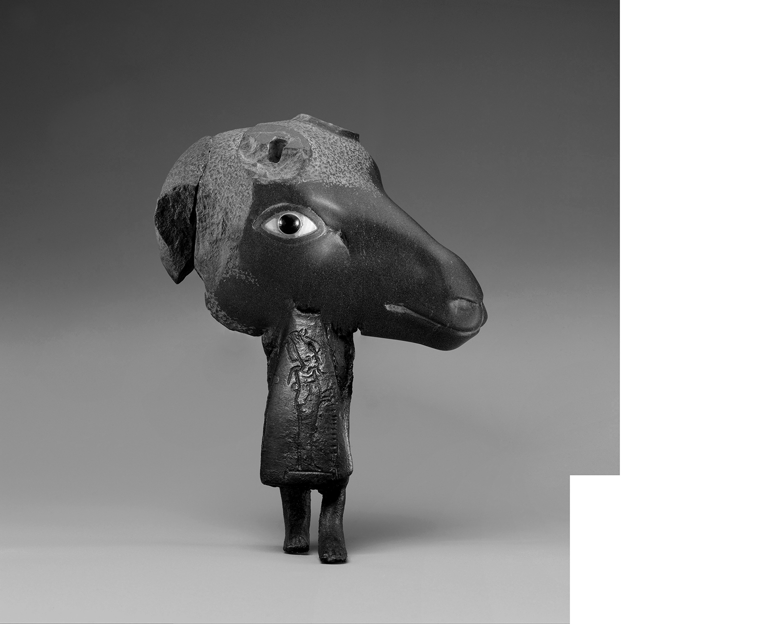 A black and white stone animal bust reminiscent of a sheep/goat. It rests on a small humanoid body in a dress, with only feet and legs visible.