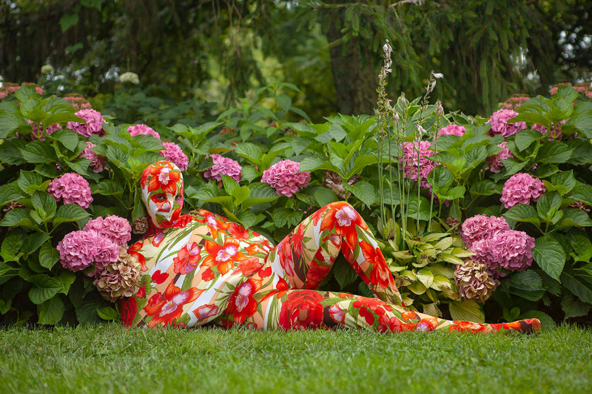 Lying in the grass among blooming rhododendrons, a figure is clad head to toe in a red, purple, and green floral ensemble that covers their body.