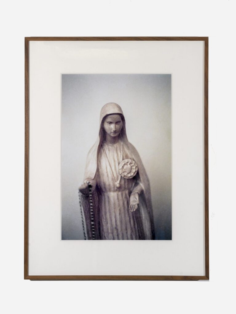 A marble statue of a woman with an outstretched left hand and prayer beads in her right. She wears a cloak, robe, and necklace. Over her heart is a circular symbol.