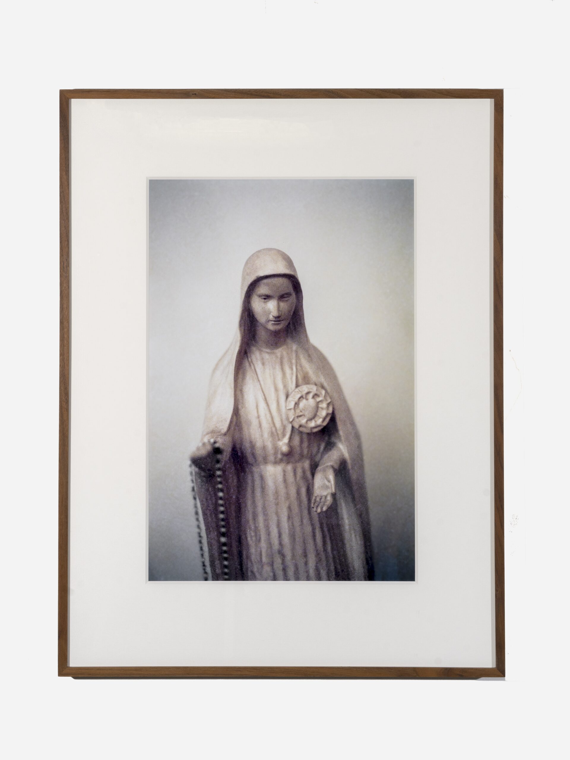 A marble statue of a woman with an outstretched left hand and prayer beads in her right. She wears a cloak, robe, and necklace. Over her heart is a circular symbol.