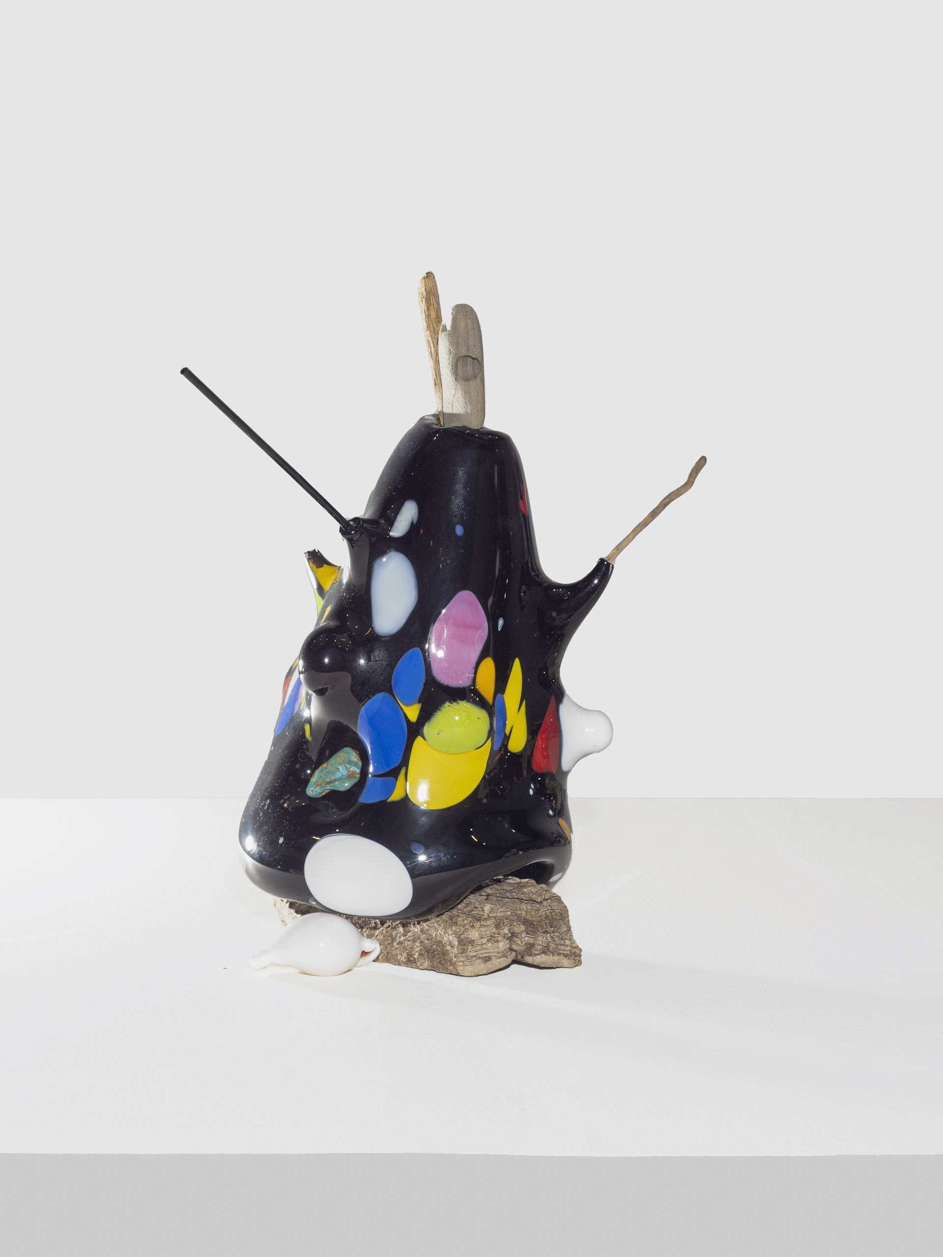 Tall glossy black sculpture with rounded form, pierced by sticks, decorated with bright oval shapes in yellow, pink, blue, white, and red, resting on rough bark and a white surface.