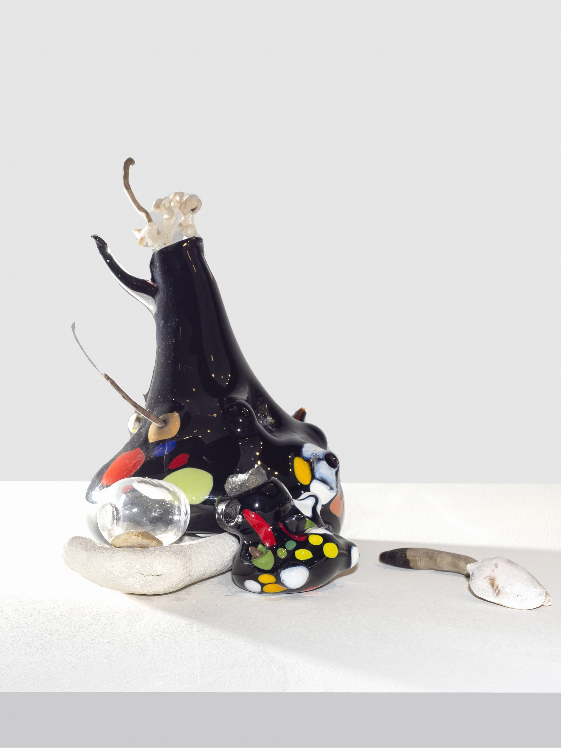Glossy black sculptural form with curving extensions and bright polka-dot shapes in red, yellow, green, and white. Next to it is a coral, glass sphere, and shell elements.