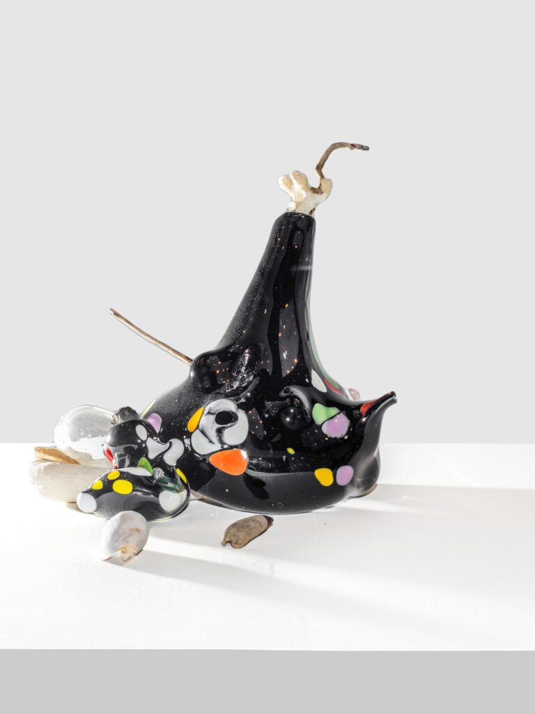 Glossy black sculpture with speckled surface and colorful dots in orange, yellow, pink, white, and green. Wood and glass elements extend from its organic form on a white platform.