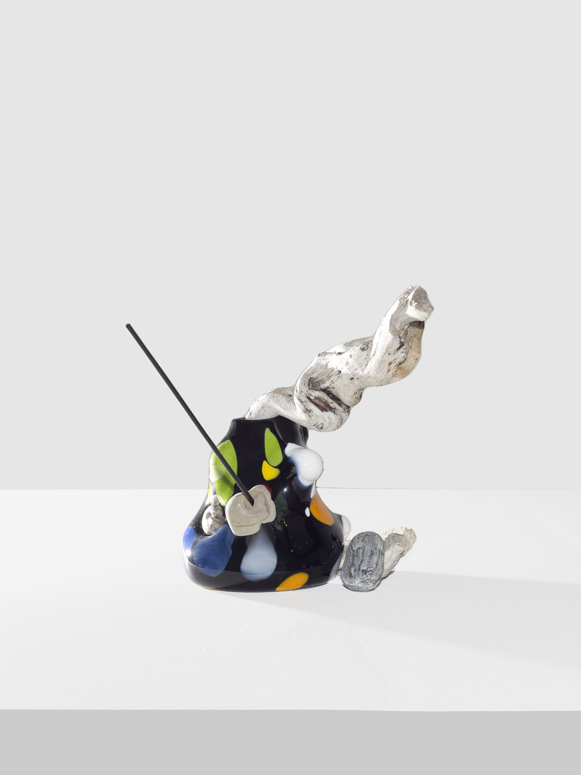 Small glossy black sculpture with bright colorful marks in green, blue, yellow, orange, and white. Wooden and metal pieces poke out from the top and base.