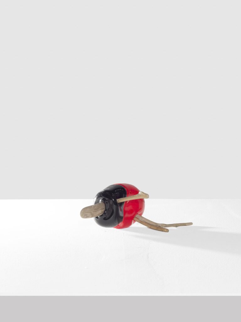 Glossy red and black elipse sculpture pierced by smooth wooden sticks, one straight and two branching, displayed on a white platform against a pale gray background.