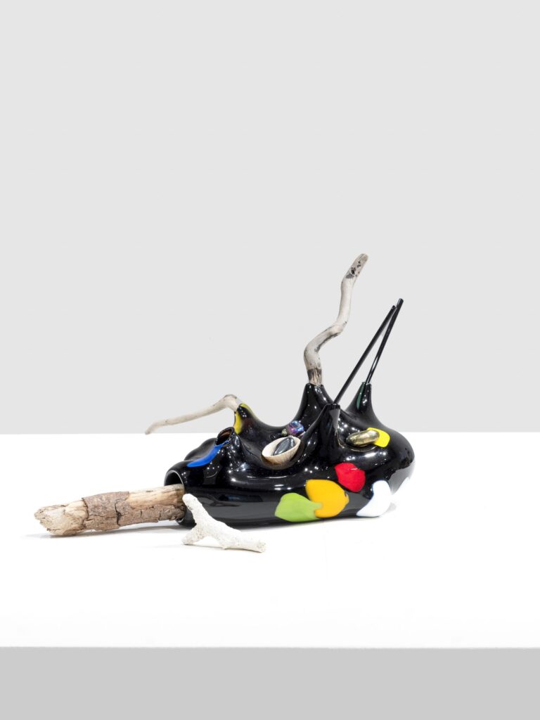 Glossy black sculpture with colorful marks in red, yellow, green, white, and blue, embedded with shells and crystals. Branches and coral elements extend from the form.