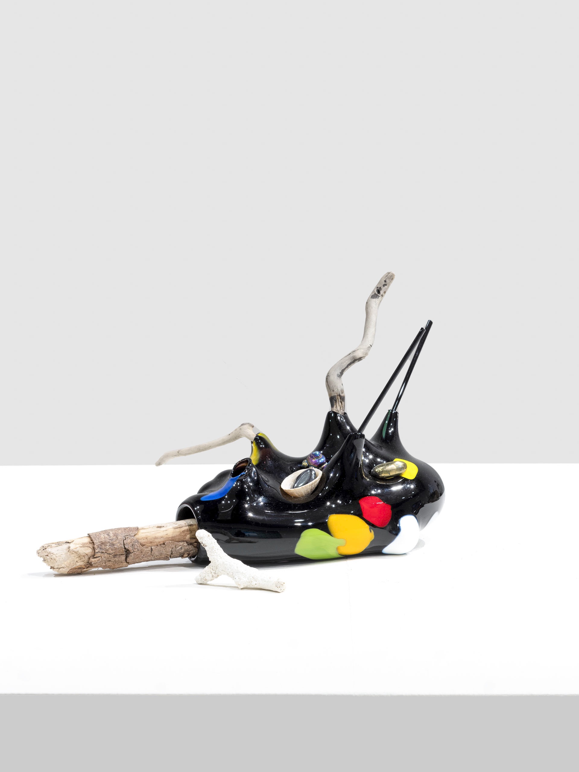 Glossy black sculpture with colorful marks in red, yellow, green, white, and blue, embedded with shells and crystals. Branches and coral elements extend from the form.