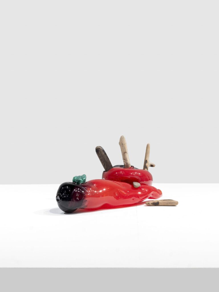 Glossy red and black glass sculpture with a melted, organic shape. Wooden sticks and a green textured stone emerge from the top, arranged on a clean white surface.