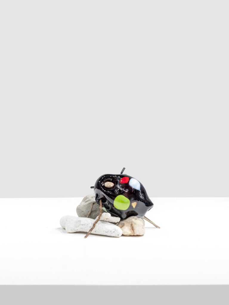 Glossy black sculptural form with colorful circular marks in red, green, blue, and beige, resting on pale stones and pierced by thin sticks, displayed on a white surface.